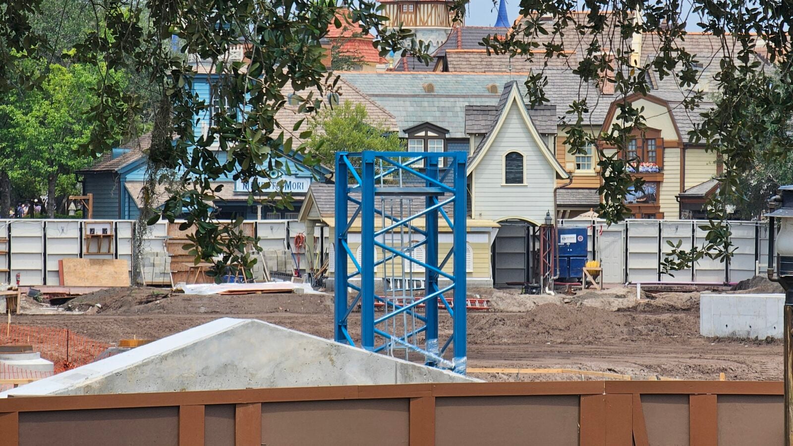 Construction site with blue steel framework in the foreground and pastel cottage style buildings in the background framed by tree branches