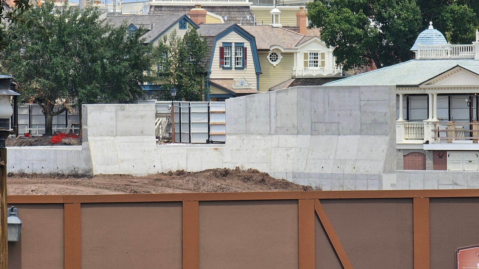 Construction site in a residential area with tall gray concrete barriers and piles of dirt in the foreground colorful houses and trees in the background