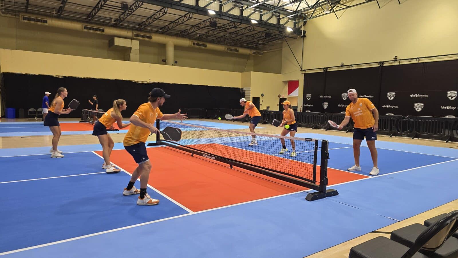Six players in orange shirts play pickleball on a blue and orange indoor court with a net in a spacious gymhall actively hitting balls and paddles ready for rallysides
