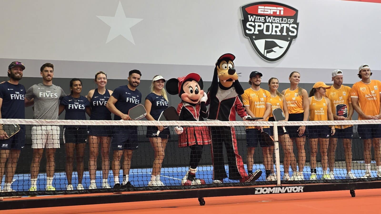 Group of athletes standing along a tennis net with mascots Minnie Mouse and Goofy ESPN Wide World of Sports sign in the background some in navy shirts reading THE FIVES and others in orange Advent Health shirts