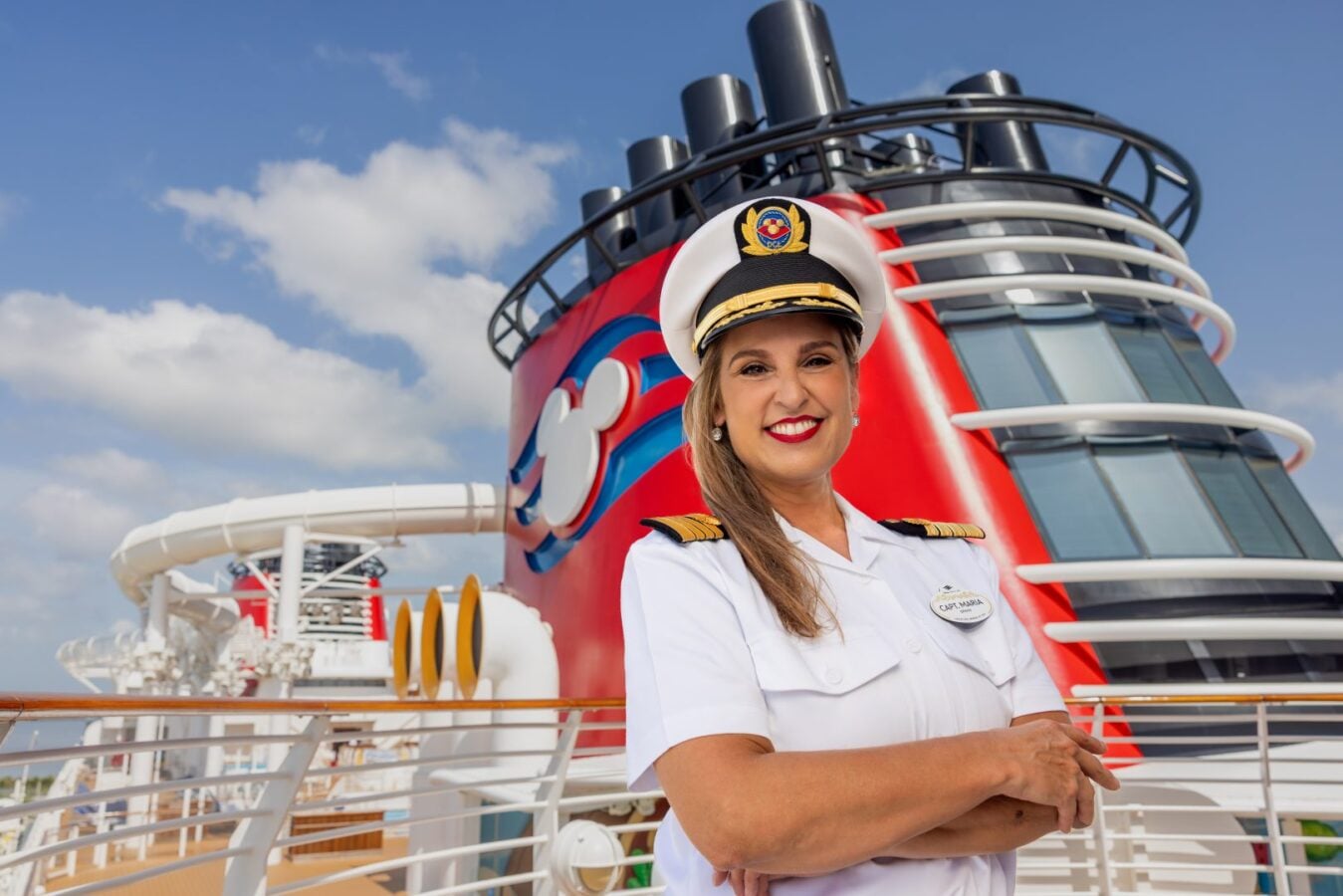 Cruise ship captain in a white uniform and hat smiling on the deck with a red ship funnel behind her