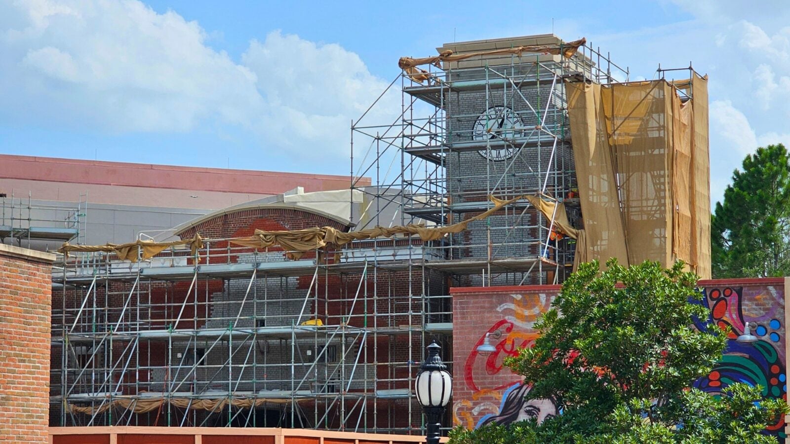 Construction site with metal scaffolding around a brick building brown protective tarps and a colorful mural along the right wall under a blue sky