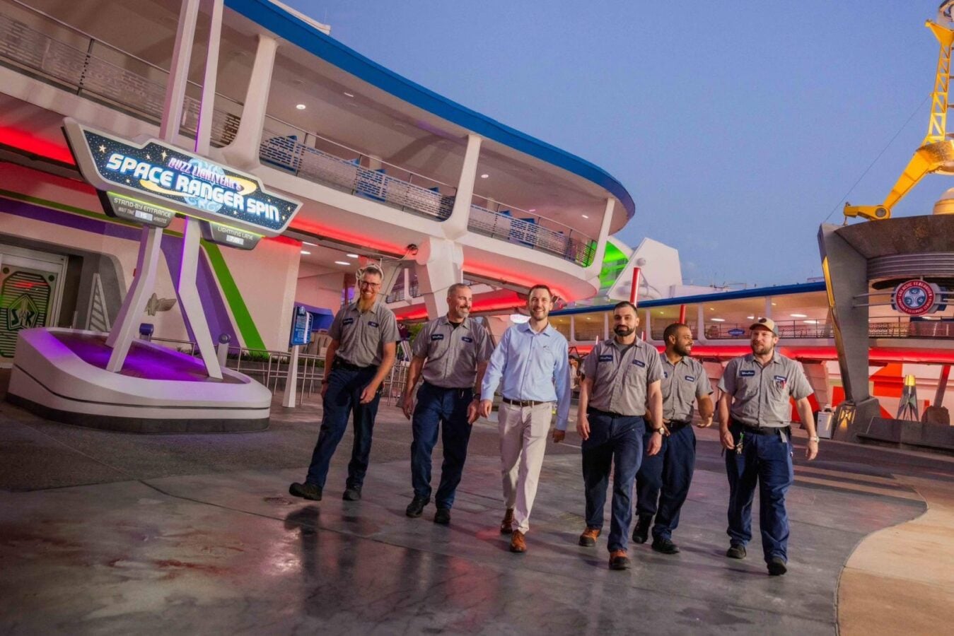 Six uniformed staff members walk toward the camera in a neon lit space themed ride area with a Space Ranger Spin sign overhead