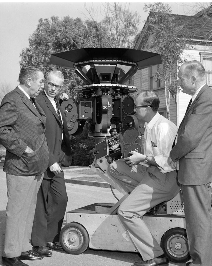 A group of men in suits inspect a large vintage film camera mounted on a wheeled platform outdoors