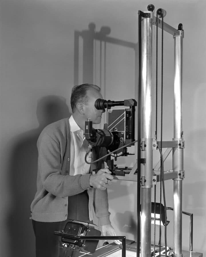Man in a cardigan uses a binocular eyepiece on a tall laboratory optical instrument at a workbench performing an alignment task