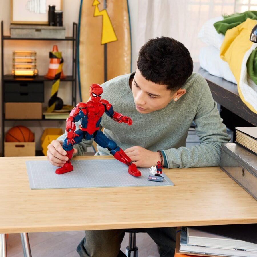 Boy at a wooden desk poses a red and blue Spider Man action figure on a gray cutting mat with small figures nearby