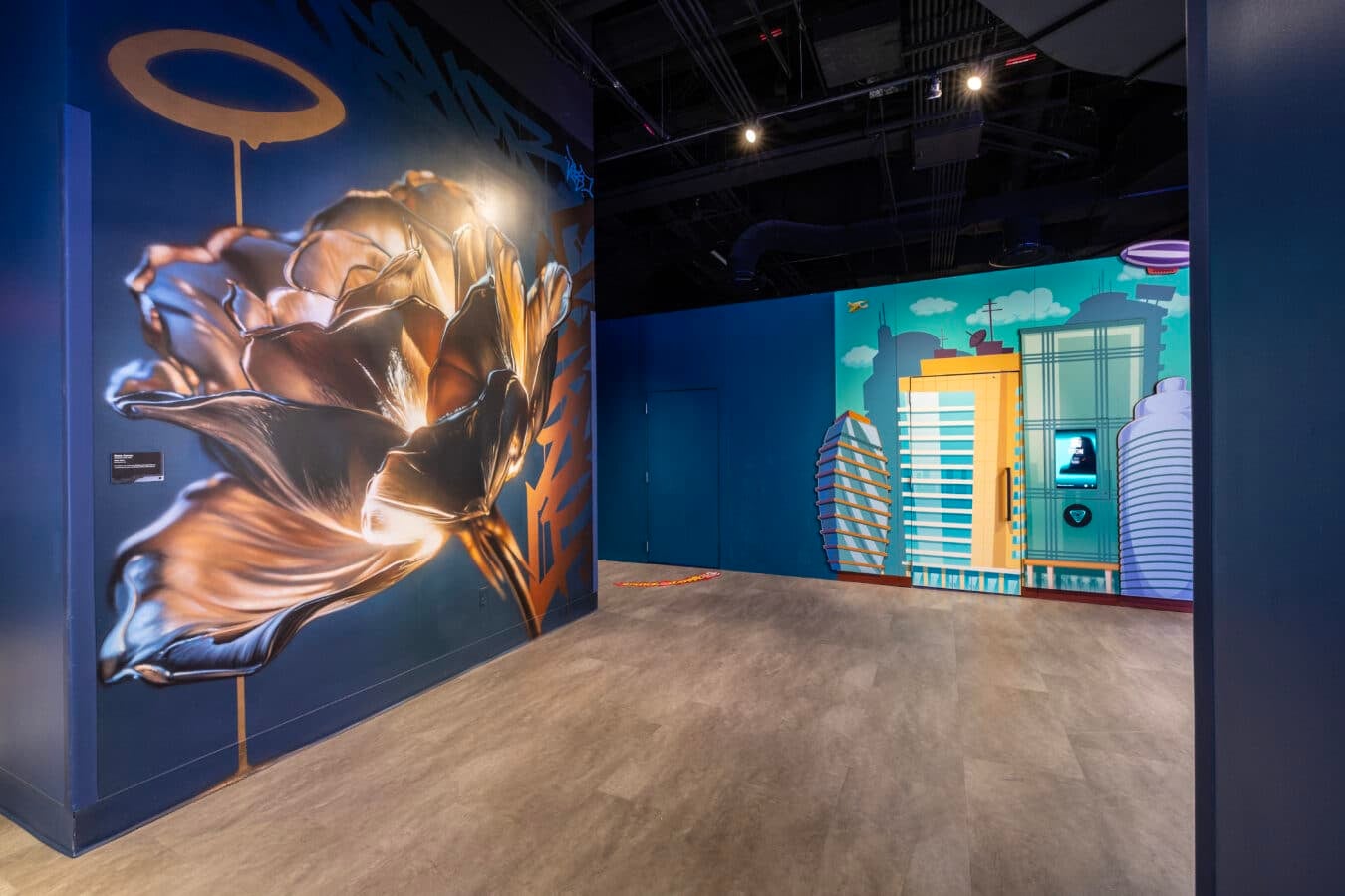 Gallery hallway with blue walls a large gold metallic looking figure mural on the left and a colorful cityscape mural on the right