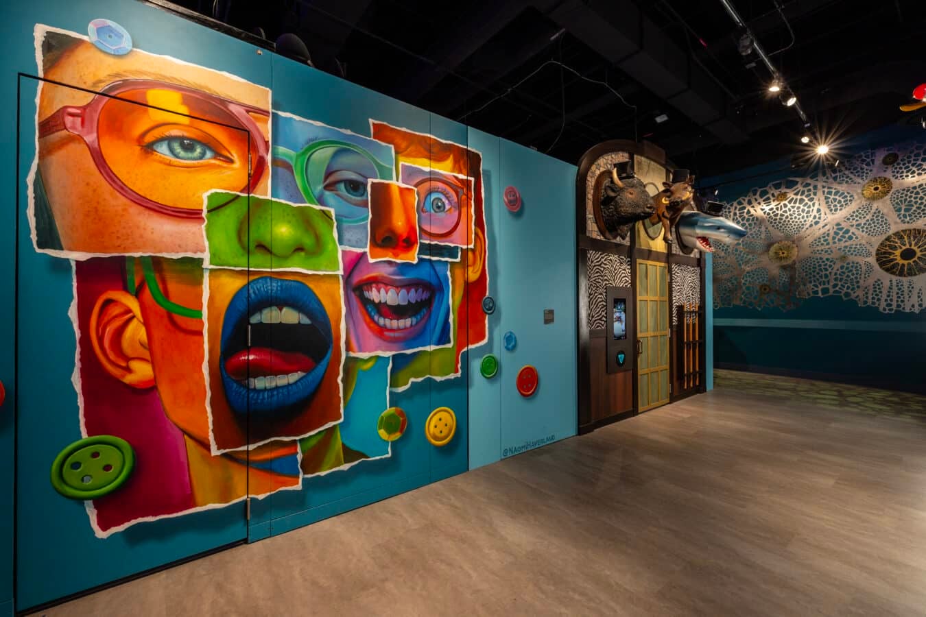 Colorful collage style mural of fragmented faces on a blue gallery wall with oversized buttons inside an exhibit space