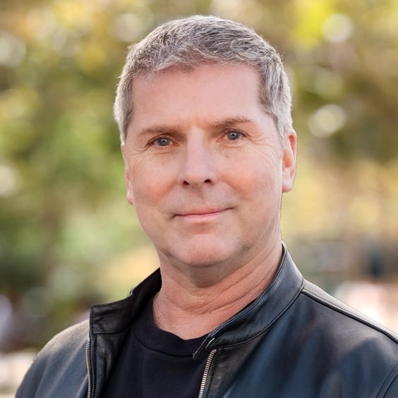 Portrait of a man with short gray hair wearing a black jacket outdoors in a sunny park setting