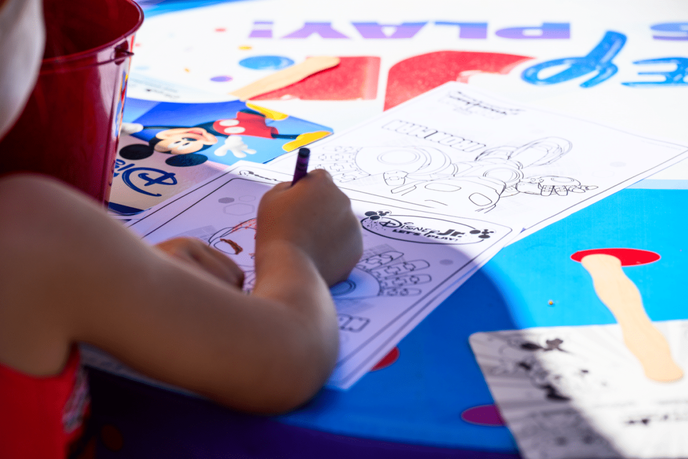 Child coloring a Disney themed page in a coloring book at a bright blue table using a purple crayon