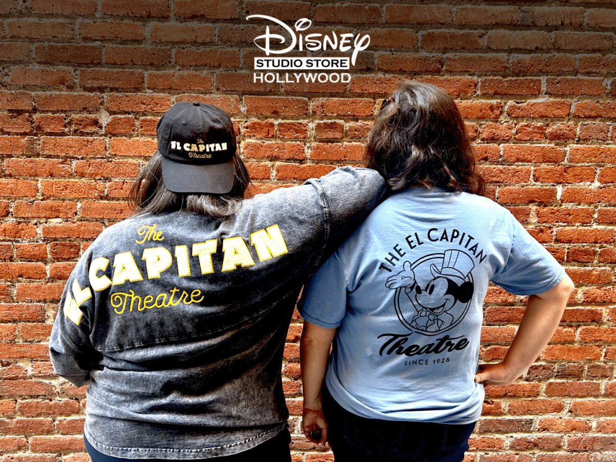 Two people with their backs to the camera wear The Captain Theatre shirts arms around each other in front of a brick wall with a Disney Studio Store Hollywood logo above
