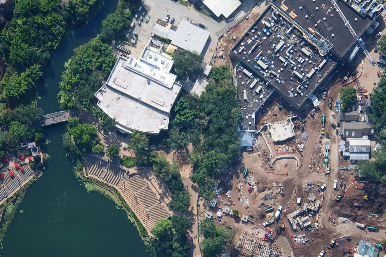 Aerial view of a construction site beside a river with cranes scaffolding and multiple buildings under development near greenery