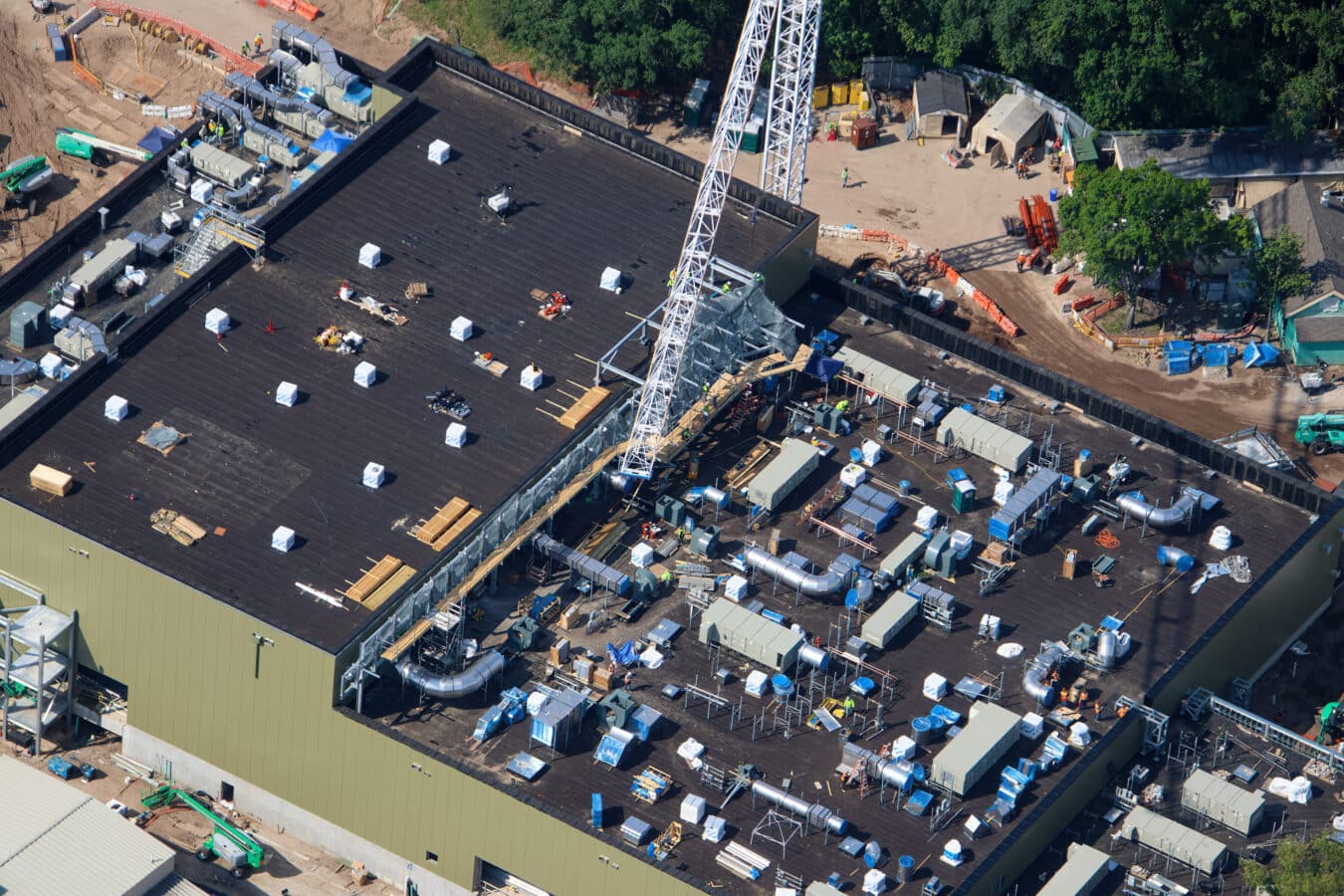 Aerial view of a construction site with a large black roof a tall crane equipment and workers around temporary structures and materials scattered across the area
