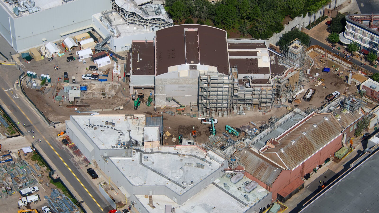 Aerial view of a large construction site with concrete buildings scaffolding machinery and white tents surrounded by roads and greenery