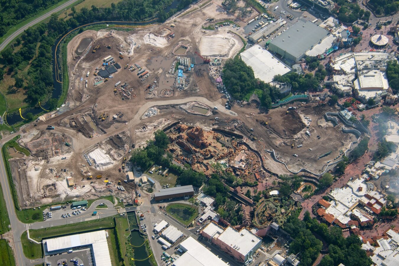 Aerial view of a large construction site with dirt piles machinery and storage areas beside a building complex and greenery