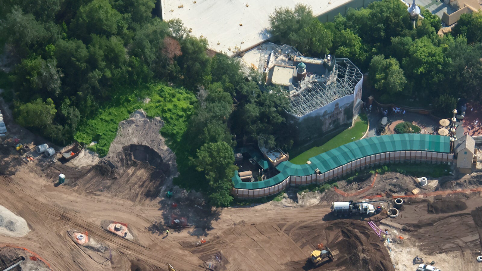Aerial view of a construction site with exposed dirt heavy equipment and a curved green roofed structure amid trees