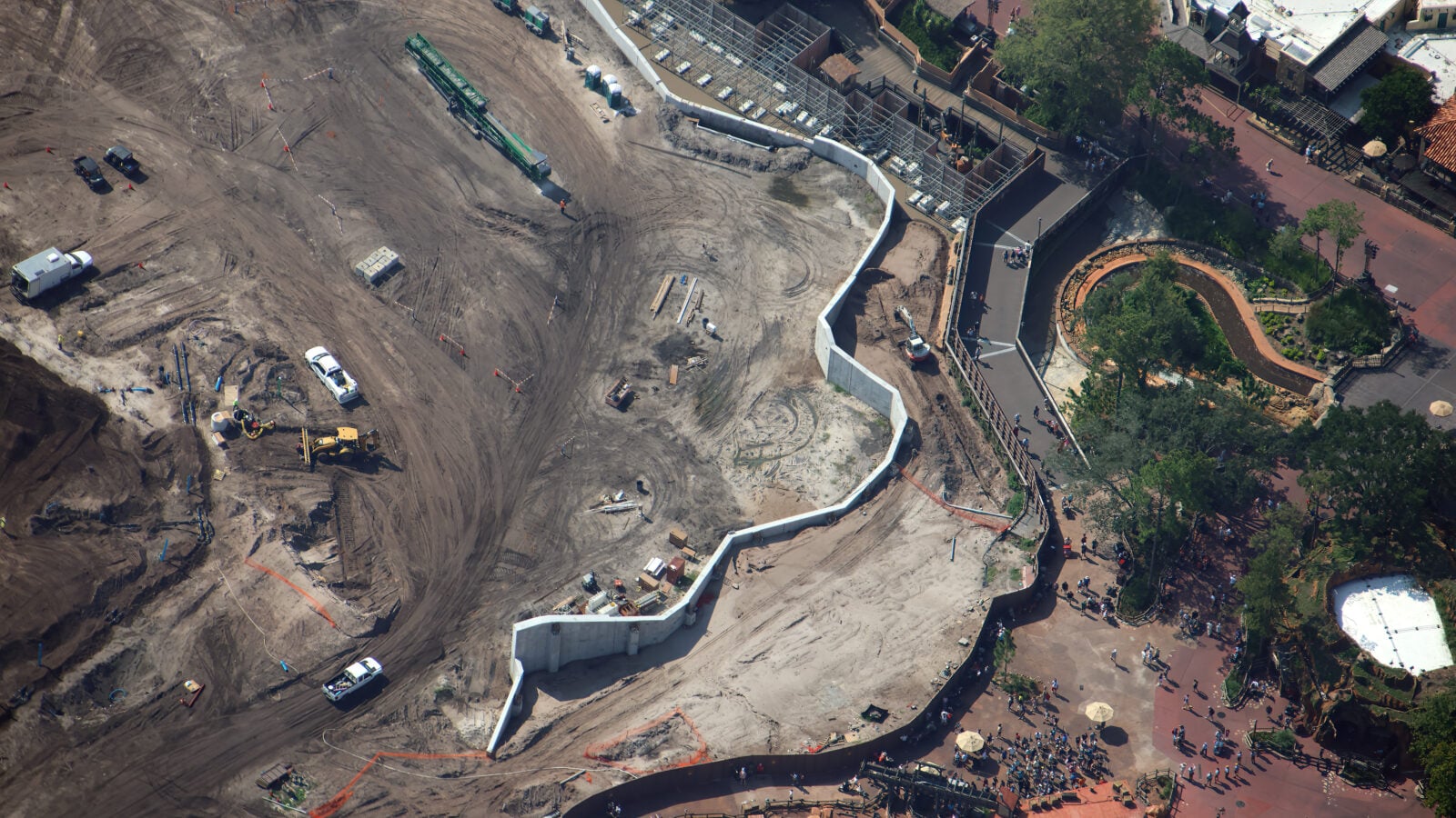 Aerial view of a construction site with dirt ground equipment and a curved white barrier along a walkway beside a plaza