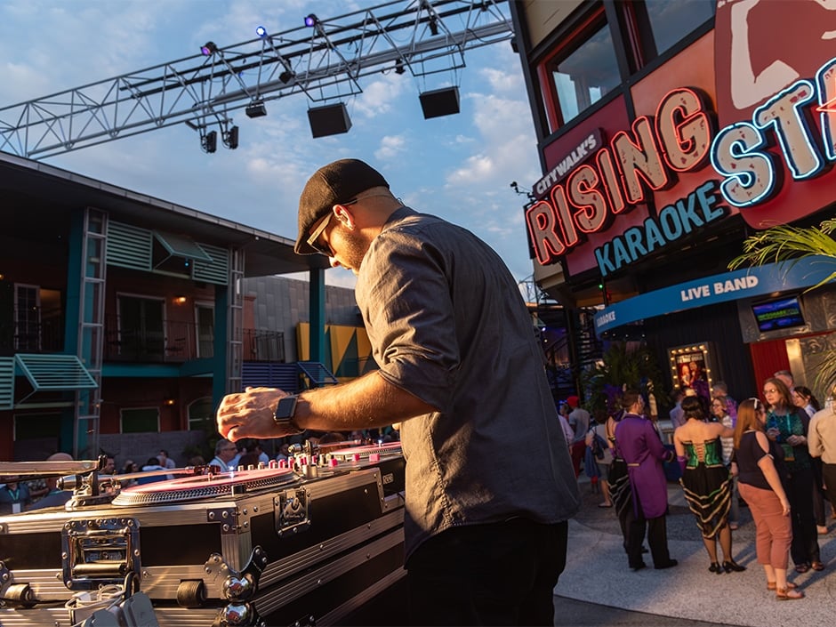 DJ playing vinyl on a booth outdoors as a crowd gathers near a neon lit karaoke bar sign that reads Rising Star