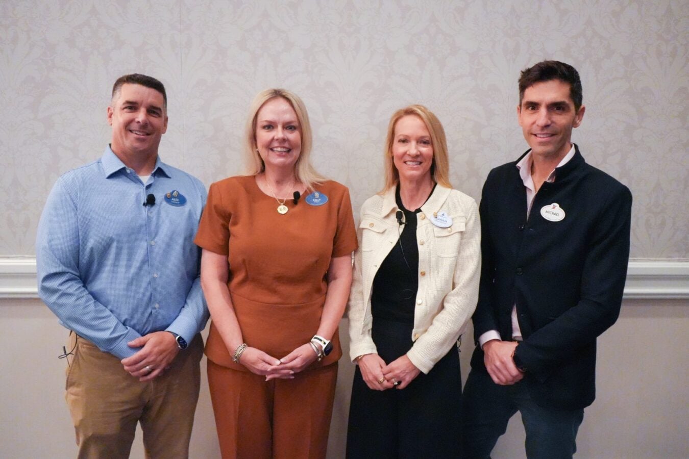 Four professionals pose together indoors against a patterned wall each wearing a name badge and a lavalier mic