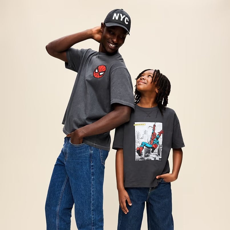 Man and boy pose in matching dark gray Spider Man Tshirts against a beige backdrop both smiling at each other