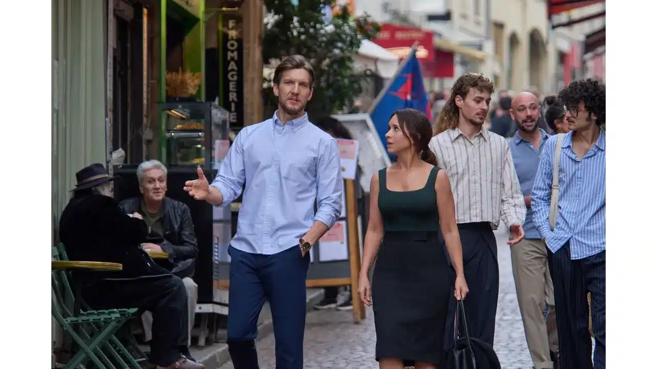 Say Oui To Romance in Paris Is Always a Good Idea on Hallmark+ Review6 Fantasy Land News Man in a light blue shirt gestures as he walks beside a woman in a dark green top and black skirt along a cobblestone street with onlookers nearby