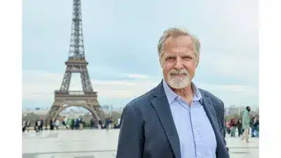 Say Oui To Romance in Paris Is Always a Good Idea on Hallmark+ Review8 Fantasy Land News Man with gray hair and beard in a blue blazer stands in front of the Eiffel Tower in Paris smiling at the camera