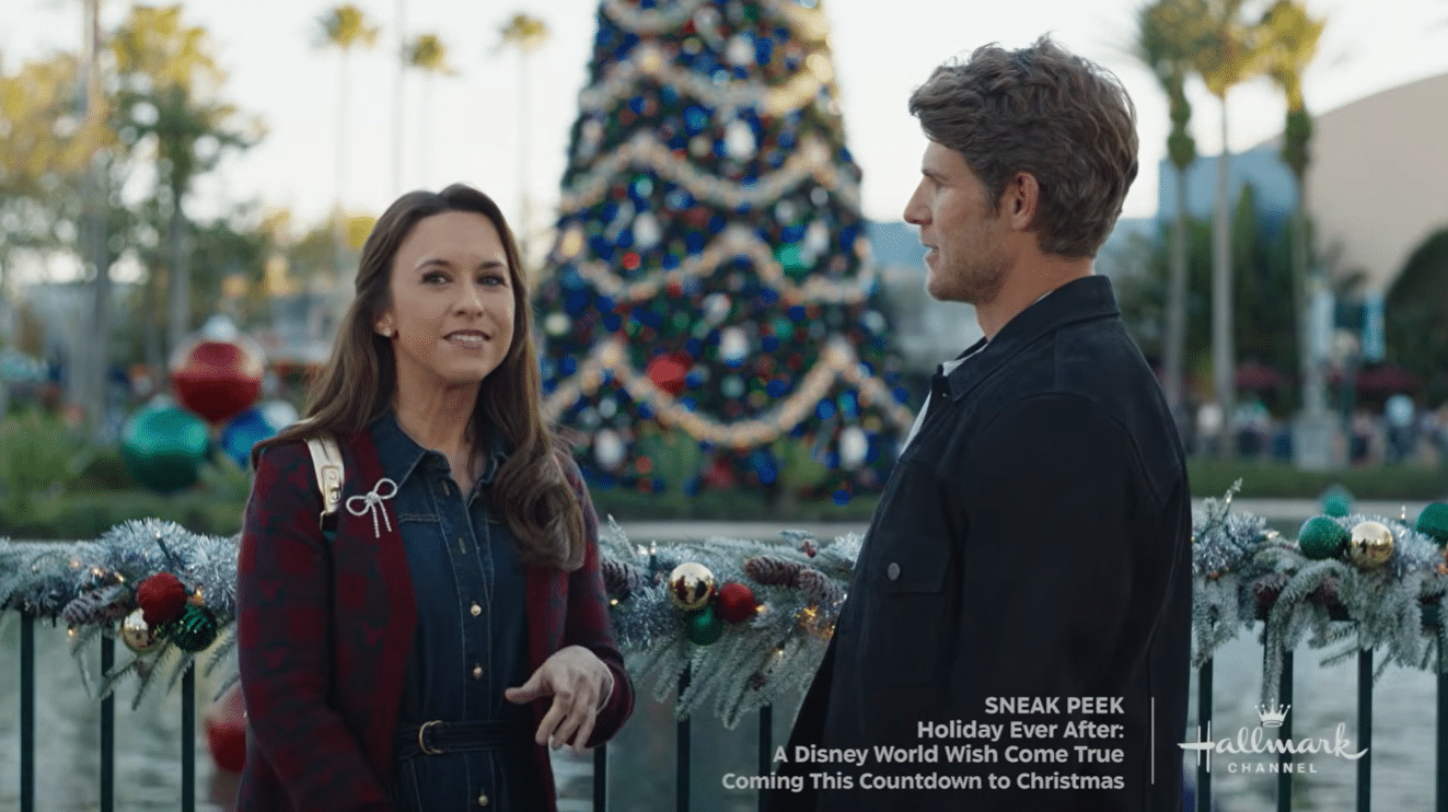 Two adults stand by a garlanded railing with a large decorated Christmas tree behind them talking outdoors Hallmark Channel watermark