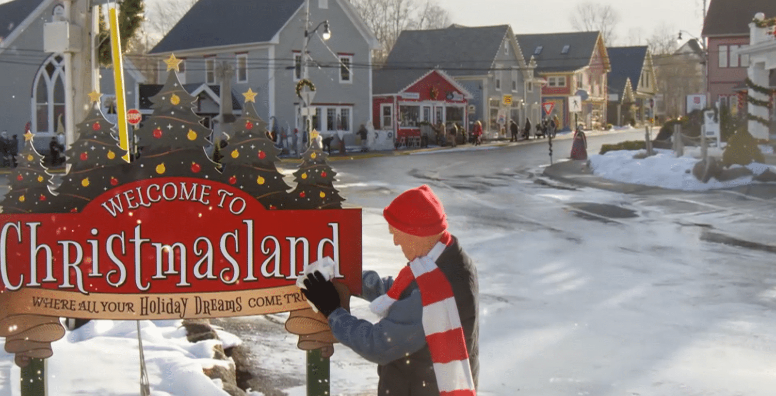 Person in a red hat and scarf stands beside a red Welcome to Christmasland sign with decorative trees on a snowy town street