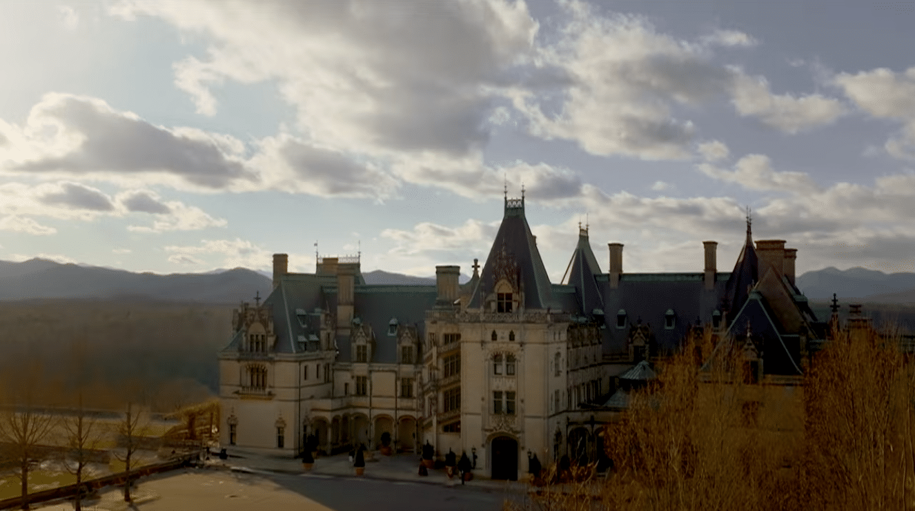 A grand European castle with blue slate roofs and tall towers set in a valley with visitors near the entrance