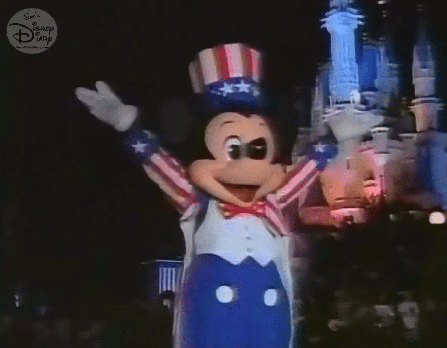 Mickey Mouse dressed in red white and blue Uncle Sam attire waving at night with a lit castle in the background