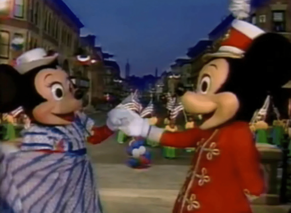 Mickey Mouse and Minnie Mouse in a parade on a city street shaking hands as cheering crowds watch