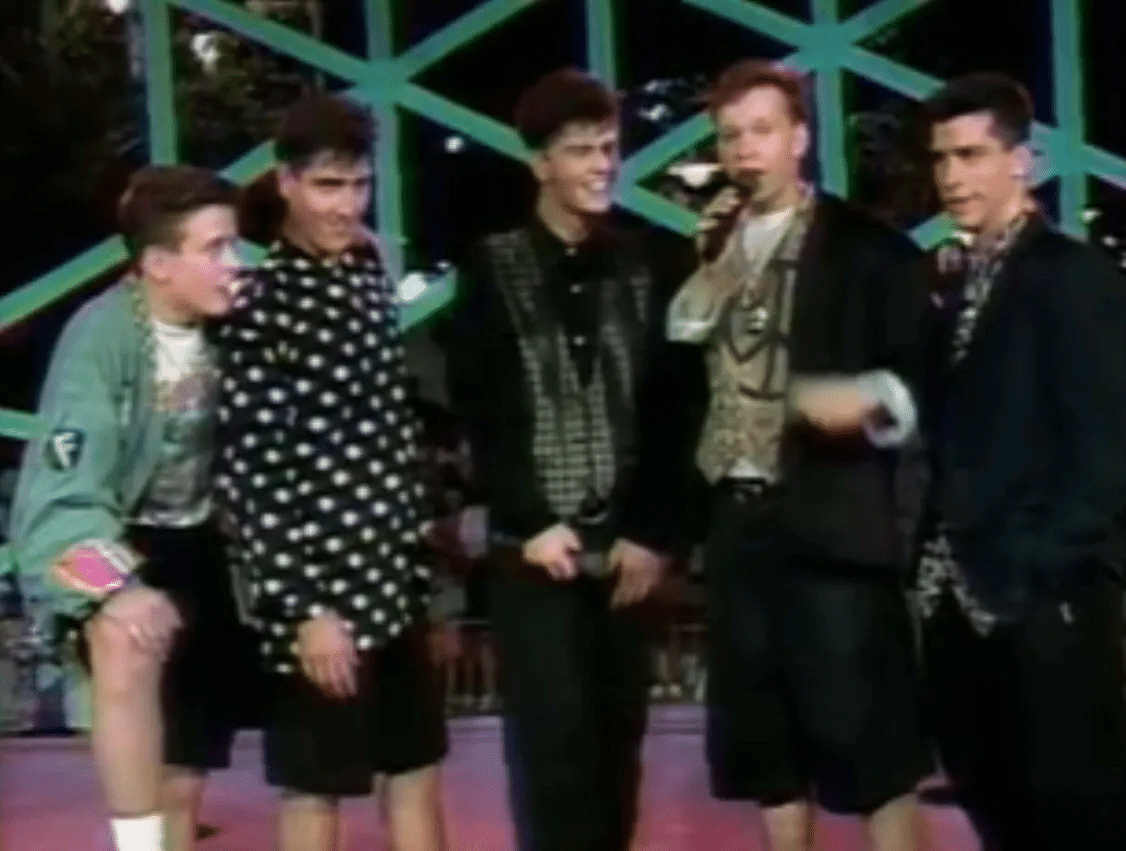 Five young men stand together on a neon lit stage one holding a microphone and speaking to an audience