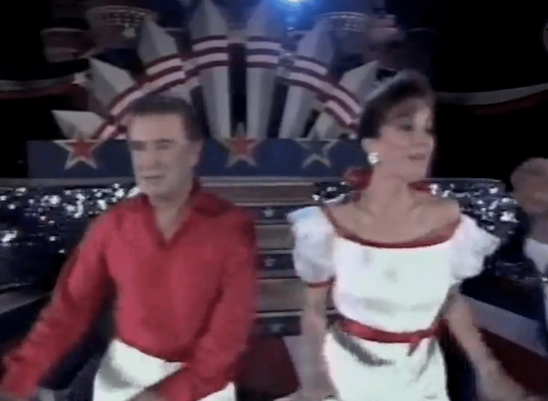 A man in a red shirt and a woman in a white dress with red trim perform on a patriotic stage backdrop with stars