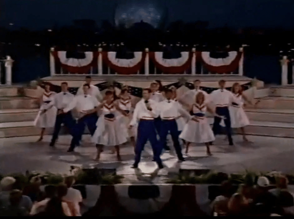 Group of dancers in white outfits and blue pants performing on an outdoor stage with red white and blue bunting in the background
