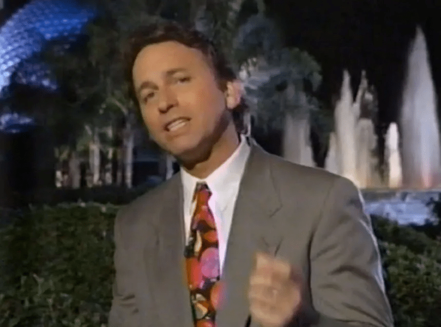 Male presenter in a gray suit with a colorful tie speaking into a microphone outdoors at night with fountains in the background