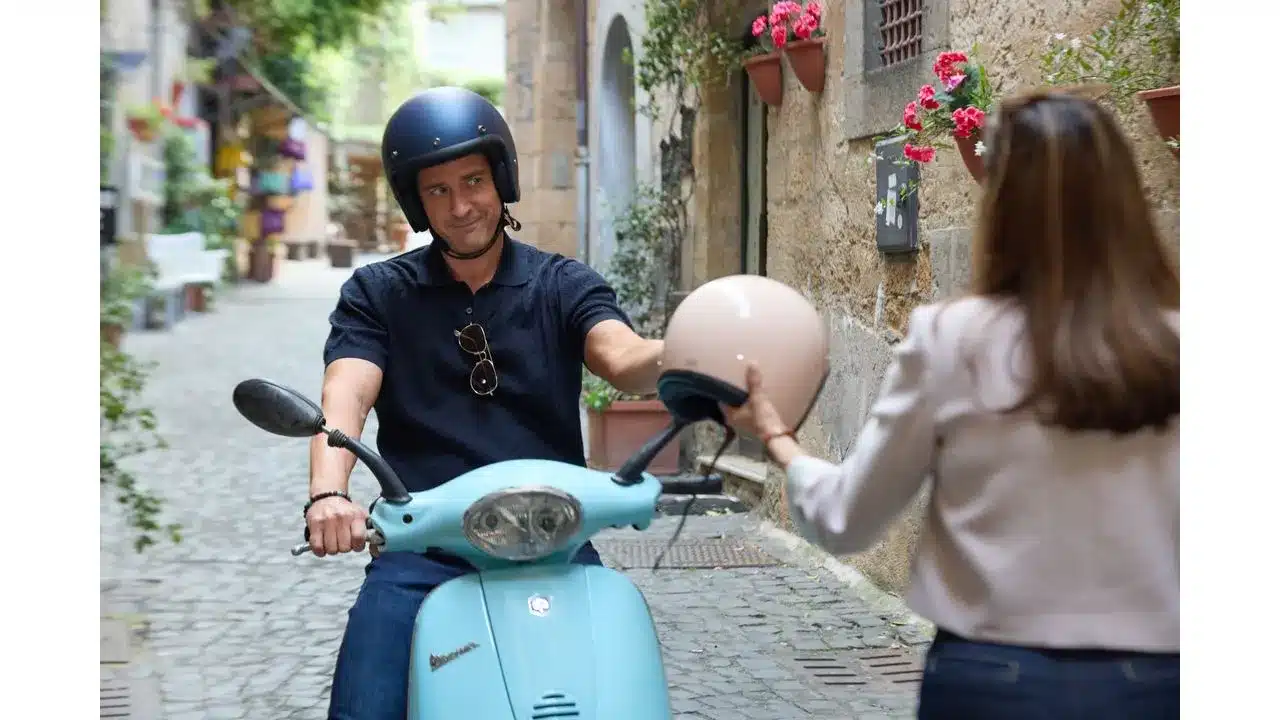 Man on a light blue scooter hands a beige helmet to a woman in a narrow stone alley with flowering pots on the wallAdmiring exchange before ride in a quaint street