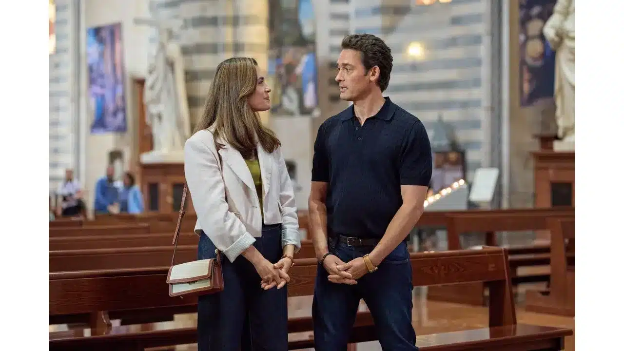 Two casually dressed adults stand in a church nave conversing beside wooden pews