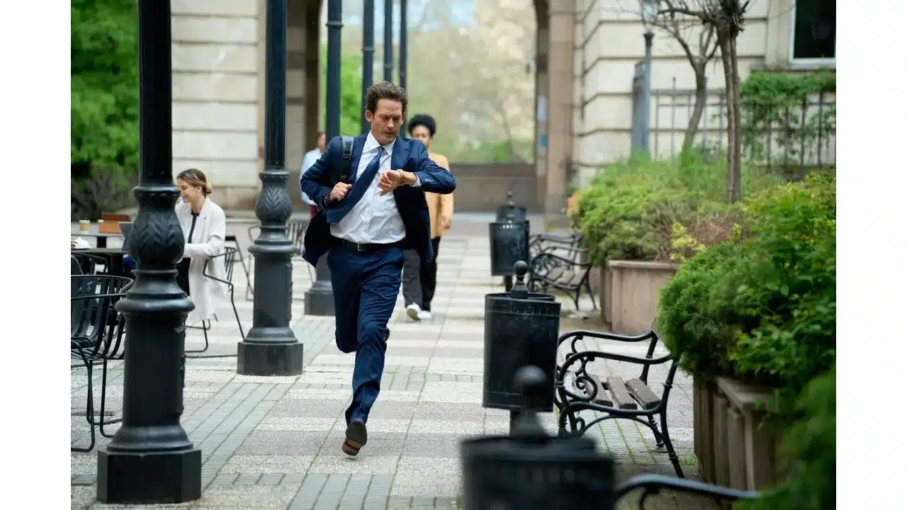 Man in a blue suit runs along a city sidewalk glancing at his watch as he hurries to a meeting