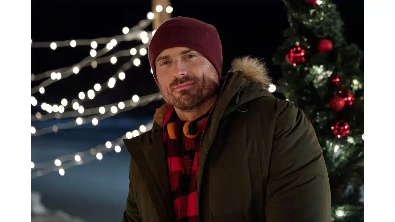 snowbound1 Fantasy Land News Man in a green parka and maroon beanie posing outdoors at night with string lights and a decorated Christmas tree