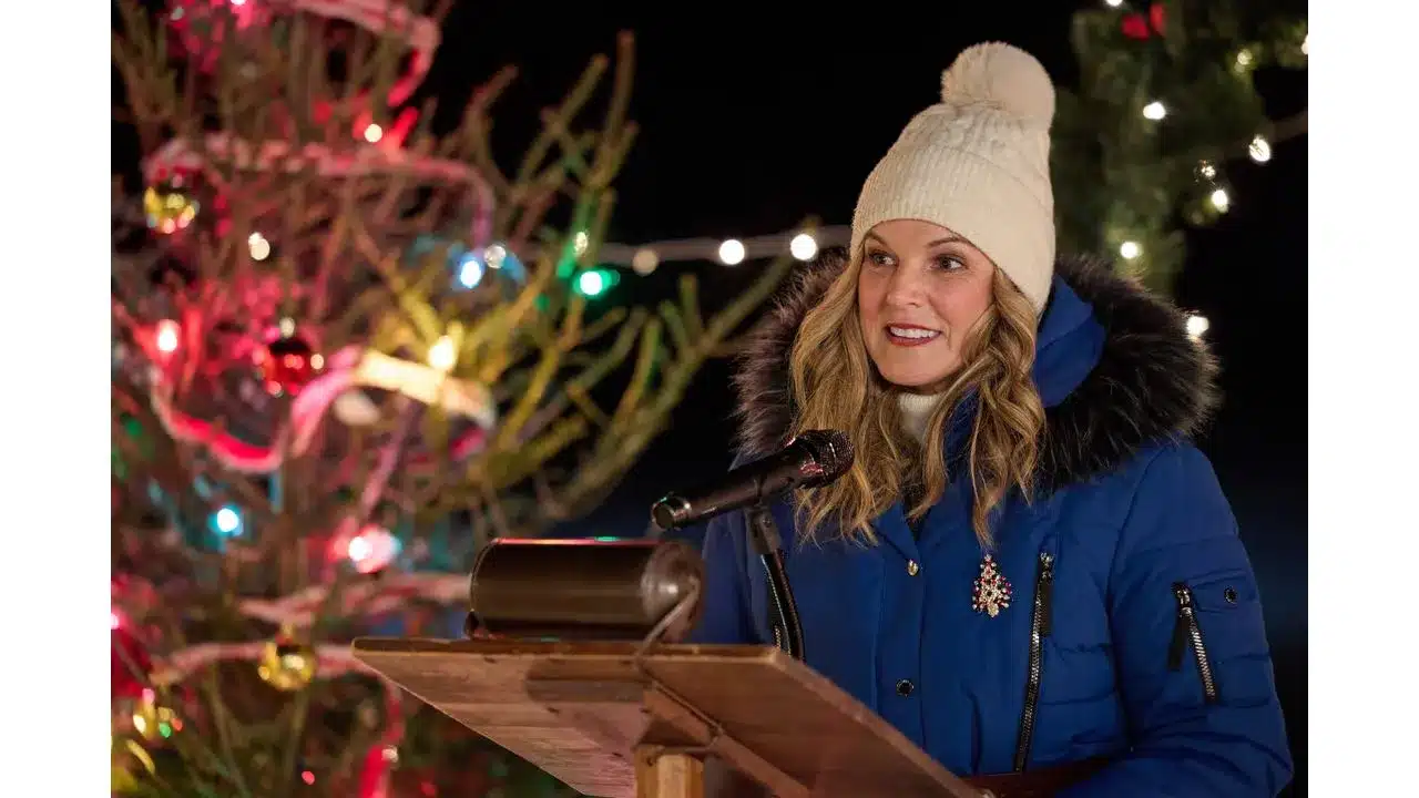 snowbound5 Fantasy Land News Woman in a blue parka with fur hood speaks into a microphone at a wooden podium during a Christmas outdoor event with festive lights in the background