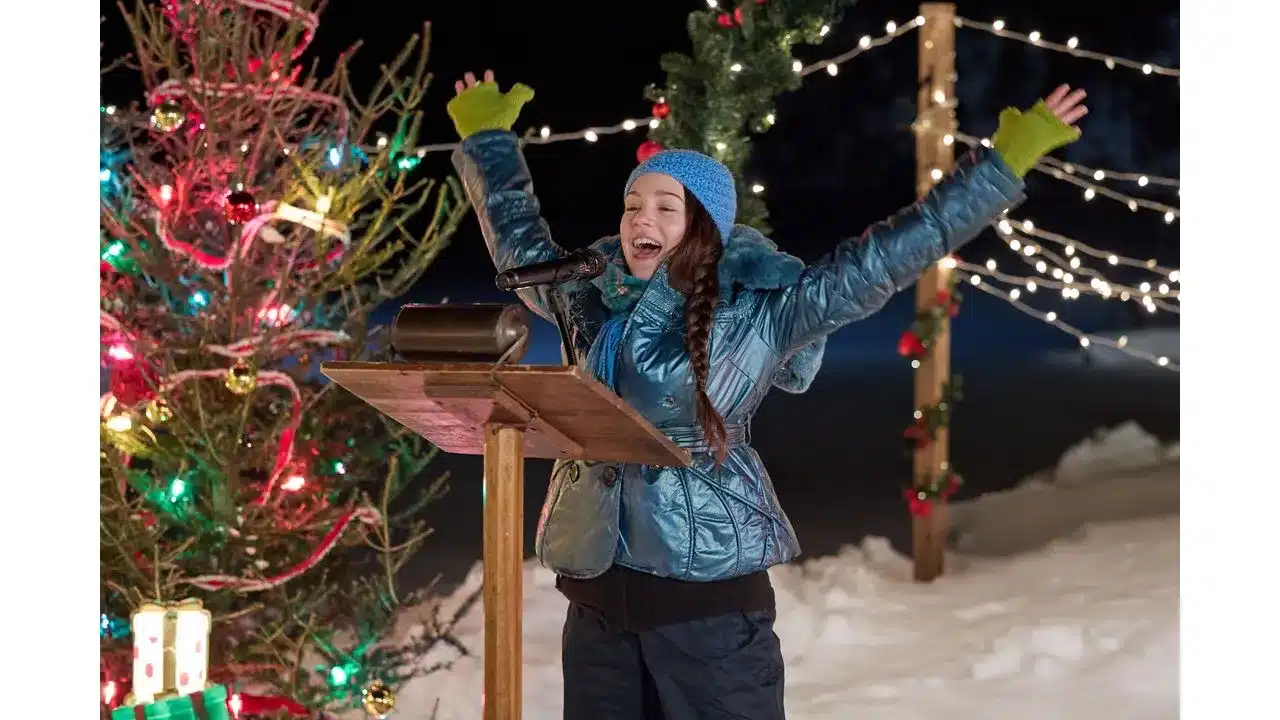 snowbound6 Fantasy Land News Young girl in a blue puffer jacket and blue knit cap raises her arms in excitement while speaking at a wooden podium outdoors at night with a Christmas tree and string lights in the snowy scene