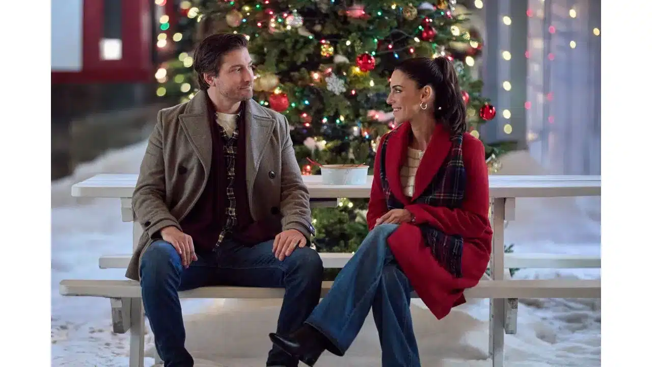 A man and woman sit on a white bench in winter chatting near a brightly decorated Christmas tree with colorful lights outside
