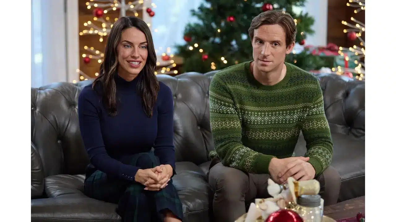 A couple sits on a gray leather sofa by a decorated Christmas tree with lights holding hands and smiling