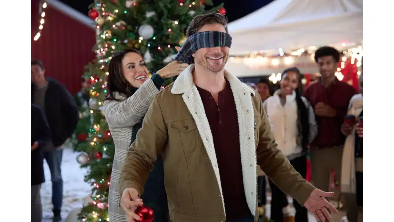 Man in a shearling lined brown jacket is blindfolded by a scarf as a woman helps him at a Christmas market with a decorated tree nearby