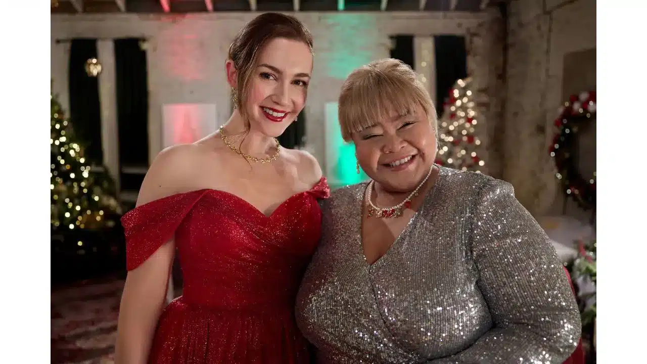 Two women in festive gowns smile for a holiday portrait with Christmas trees and colorful lights in the background
