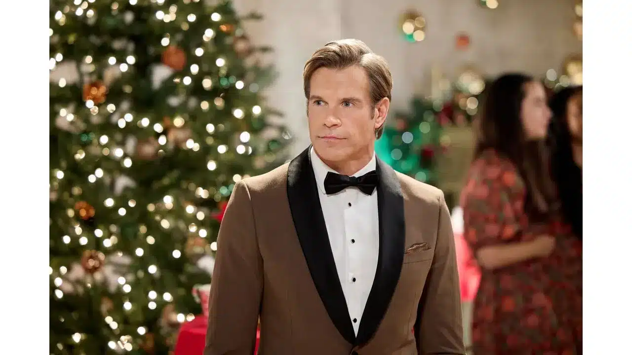 Man in a brown tuxedo with a black lapel bow tie at a holiday party with a Christmas tree and festive lights in the background