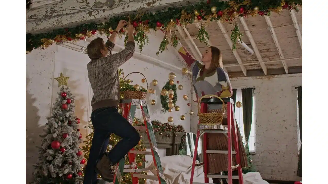 Couple on ladders decorating a rustic ceiling with garlands and gold ornaments for Christmas in a festively decorated room