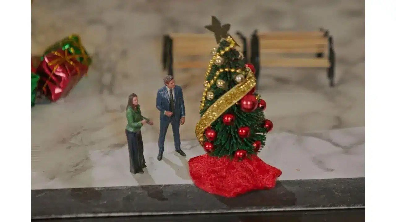 Miniature Christmas tree with red ornaments and a gold ribbon a small man in a suit and a woman in a green top stand beside it on a marble surface with a blurred bench in the background