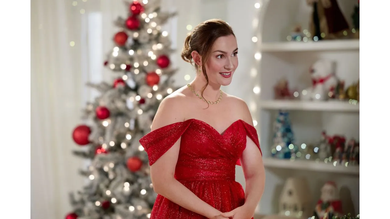 Elegant woman in a sparkling red off the shoulder gown posing indoors with a Christmas tree and twinkling lights in the background