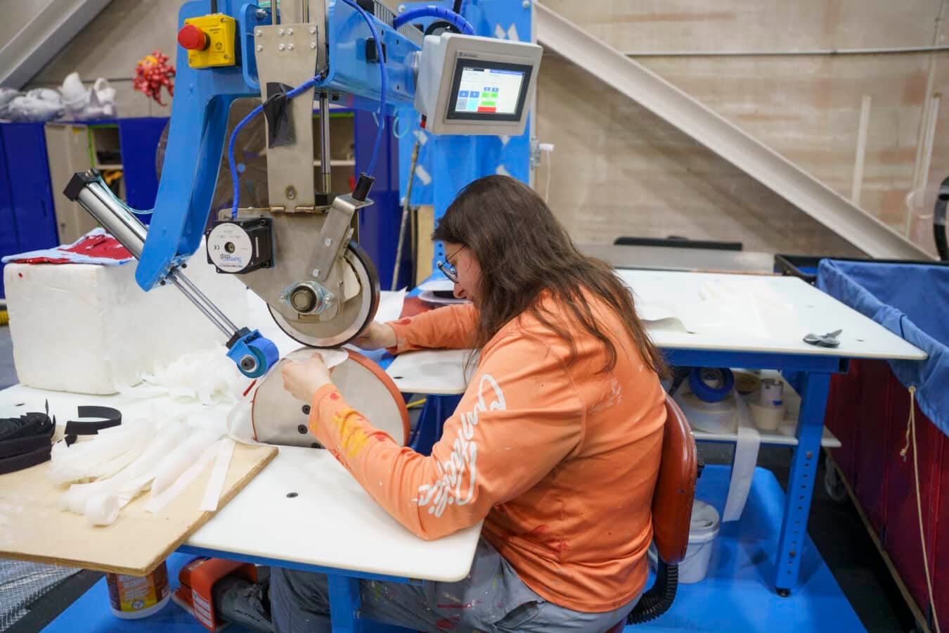 Person in an orange shirt sits at an industrial sewing machine stitching white fabric strips on a worktable